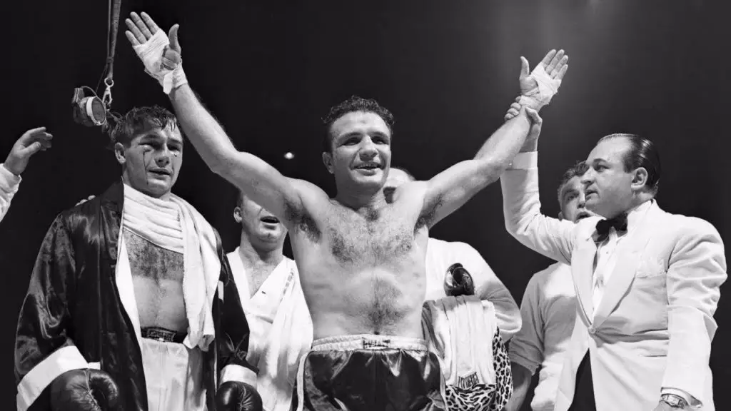 Billy Fox looks happy with his arms raised as he is pronounced the winner with Jake LaMotta looking bloodied behind him.